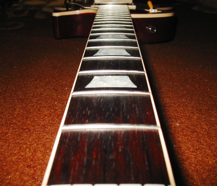 Close-up of a guitar neck with frets and inlay markers. The body of the guitar is visible in the blurred background
