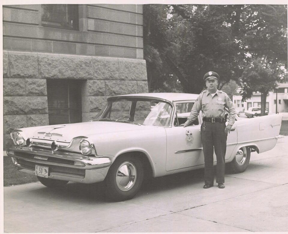 Vintage black-and-white photo of a uniformed officer beside a 1950s sedan