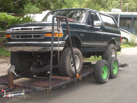 1989 Ford Bronco - Dark green 1989 Ford Bronco is loaded onto a trailer with large off-road tires.