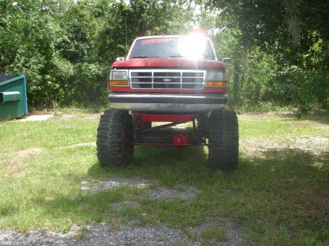 1989 Ford Bronco - Lifted red Ford truck with massive tires and a custom grille, parked outdoors.