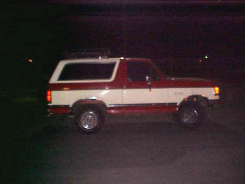1987 Ford Bronco - Side view of a two-tone red and white 1987 Ford Bronco with a roof rack, photographed at night.