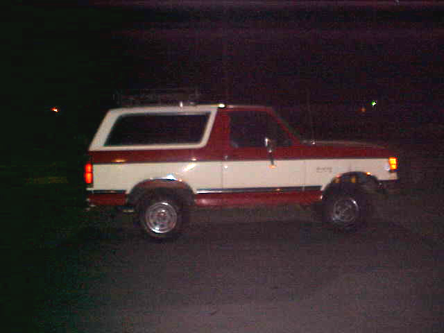 1987 Ford Bronco parked at night, side view, red and white two-tone paint, roof rack