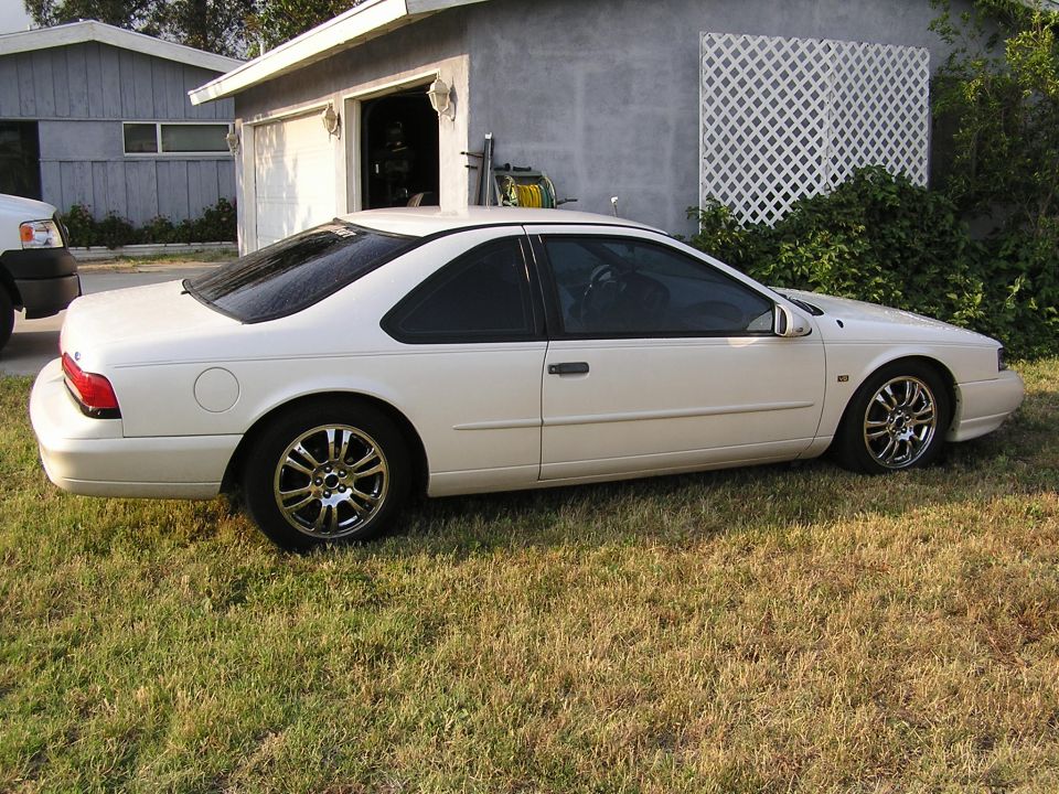 White Ford Thunderbird coupe with chrome wheels, side view, parked on grass by a gray garage