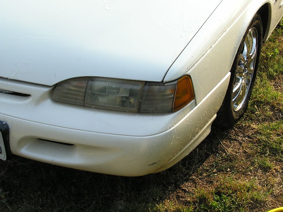 Front left corner of a white Ford Thunderbird with chrome wheel, headlight and amber marker, parked on grass