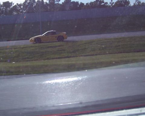 1988 Pontiac Fiero - A yellow 1988 Pontiac Fiero is seen on a race track, with a grassy embankment and trees visible.