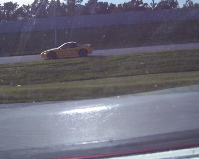 Yellow 1988 Pontiac Fiero driving on a paved track with grass and trees in the background