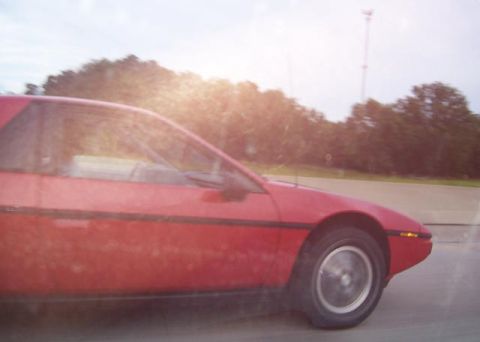 1988 Pontiac Fiero - A red Pontiac Fiero is captured in motion on a sunny day, with trees and a cell tower in the background.