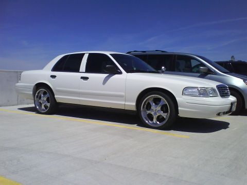 2003 Ford Crown Victoria - A white 2003 Ford Crown Victoria sedan is parked in a lot, custom chrome wheels and tinted windows.