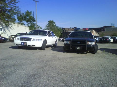 2003 Ford Crown Victoria - Two Ford Crown Victoria Police Interceptors, one white and one black with light bar, parked in a lot.