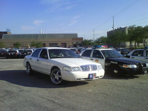2003 Ford Crown Victoria - A white 2003 Ford Crown Victoria with custom chrome wheels is parked in a lot filled with police vehicles.