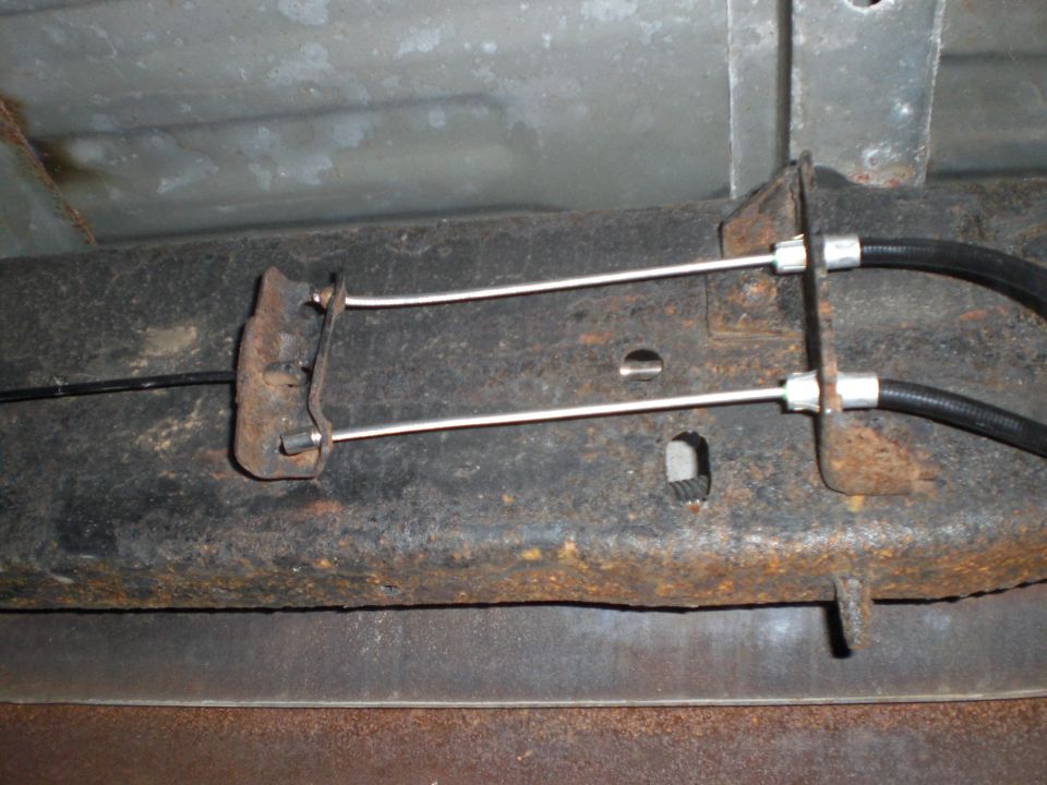 Underside of a vehicle frame with two brake cables attached by rusty brackets.