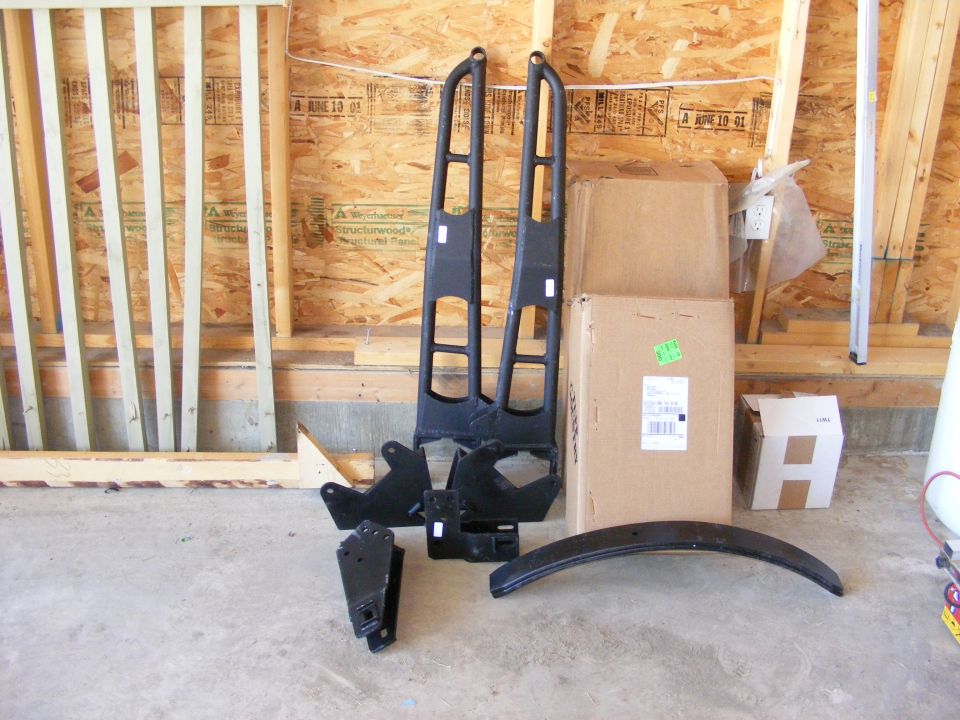 Truck lift kit parts laid out on a concrete floor in front of a wooden wall. Includes two tall black metal pieces, brackets, and a curved black piece