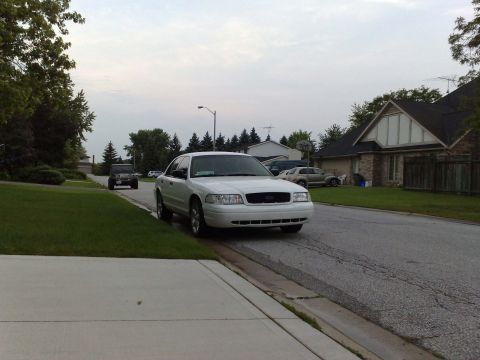2003 Ford Crown Victoria - A white Ford Crown Victoria sedan is parked on the right side of a residential street, facing the camera. A black Jeep Wrangler is parked f…