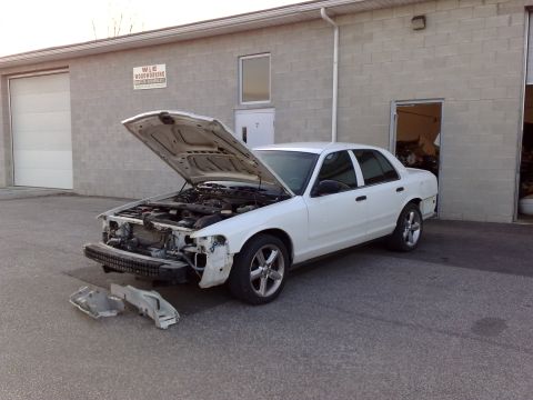2003 Ford Crown Victoria - A white sedan with its hood open is parked in front of a brick building. The front bumper and some other parts are removed and lying on the…
