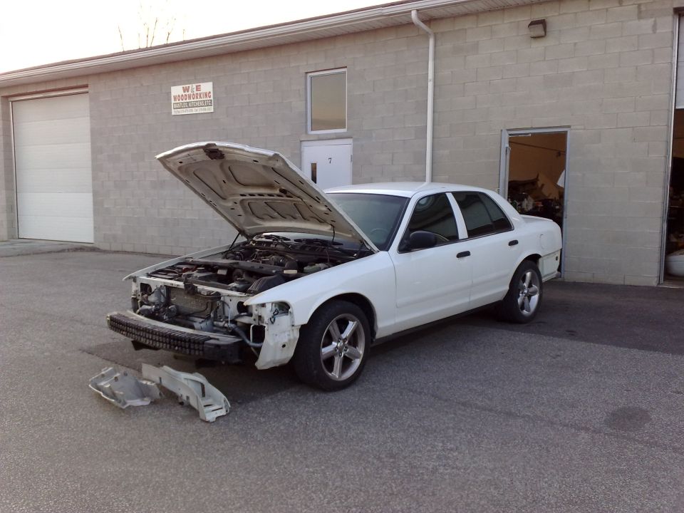 White sedan with hood open, front bumper removed, parked outside a building.