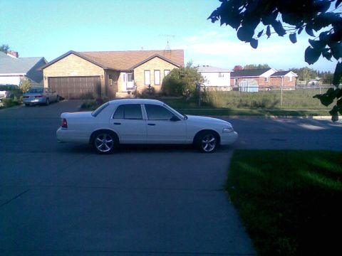 2003 Ford Crown Victoria - A white 2003 Ford Crown Victoria sedan is parked on a paved driveway in front of a brick house. The car is viewed from the side, showing it…
