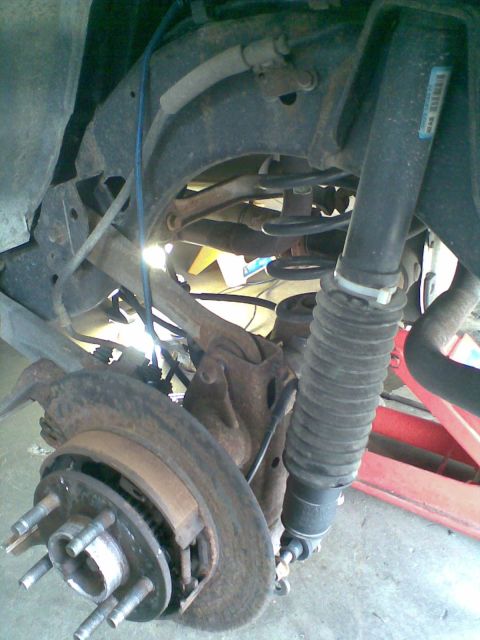 2003 Ford Crown Victoria - Close-up of a car's front corner with the wheel removed, exposing a rusty vented brake rotor, hub with five wheel studs, caliper bracket an…