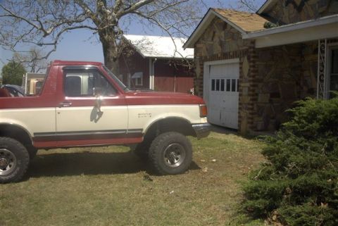 1987 Ford Bronco - This lifted 1987 Ford Bronco features a two-tone red and white paint scheme and is parked in a grassy yard next to a garage.