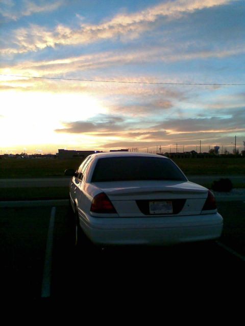 2003 Ford Crown Victoria - White Ford Crown Victoria sedan shot from directly behind in an empty parking lot at sunset, backlit by an orange and blue sky. The car has…