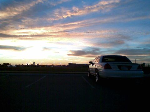 2003 Ford Crown Victoria - A white sedan is parked in a parking lot with a dramatic sunset sky in the background. The car is viewed from the rear quarter angle.