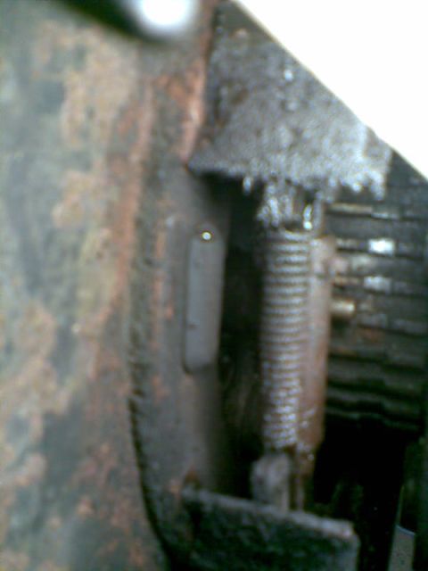 2003 Ford Crown Victoria - Blurry close-up inside a vehicle's front-end structure showing heavy surface rust and road grime on a stamped-steel panel next to a coiled…