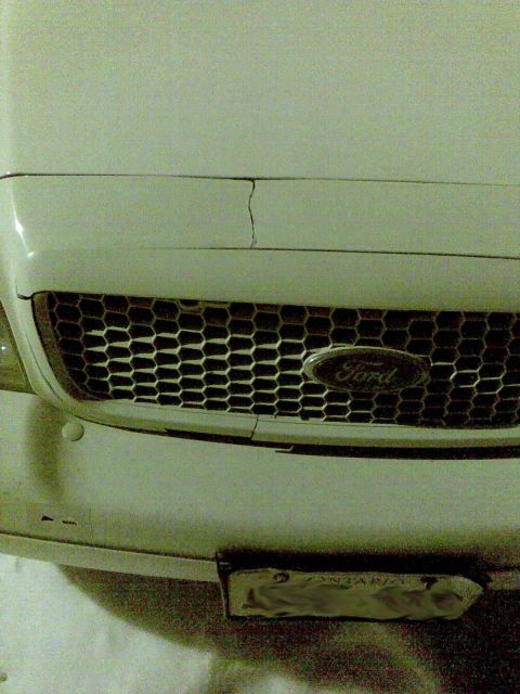 2003 Ford Crown Victoria - Grainy low-angle close-up of the front end of a white Ford, showing a honeycomb mesh grille with the Ford oval badge and a visible crack ru…