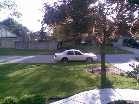 2003 Ford Crown Victoria - A white Ford Crown Victoria sedan sits at the curb of a suburban street, photographed in side profile from across a mowed front lawn with a…