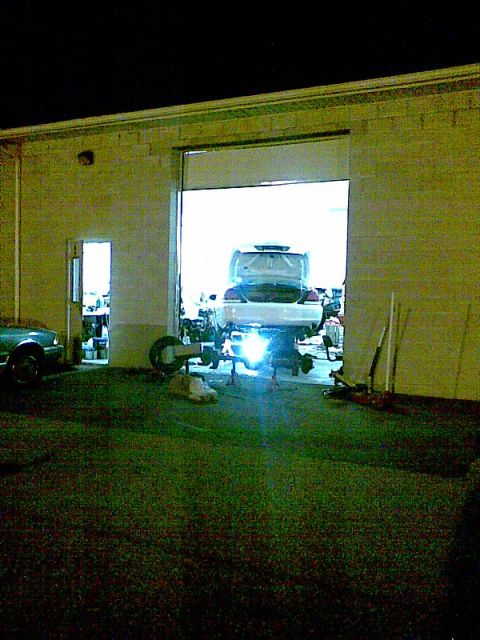 2003 Ford Crown Victoria - A white sedan is raised on a lift inside a garage with the rear end facing the camera. The garage door is open, revealing the bright interi…