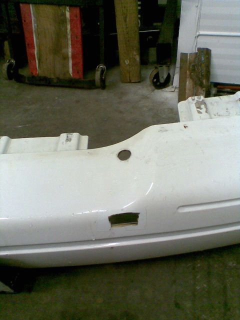 2003 Ford Crown Victoria - A white plastic rear bumper cover removed from the car and set on a shop floor, showing a round hole and a rectangular cutout in the fascia…