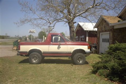 1987 Ford Bronco - Side view of a two-tone 1987 Ford Bronco parked in front of a rural building.