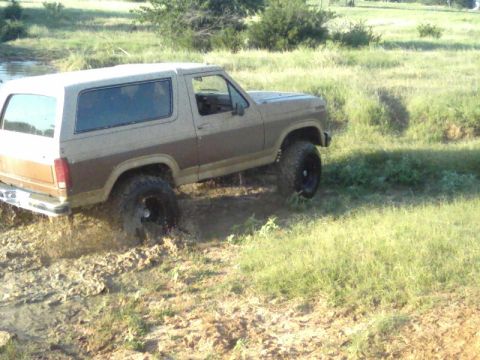 1986 Ford Bronco - This Ford Bronco is navigating a muddy off-road trail, with its tires partially submerged in the mire.