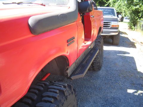 1989 Ford Bronco - A red off-road vehicle with a snorkel is parked next to a gray Ford truck on a gravel road.