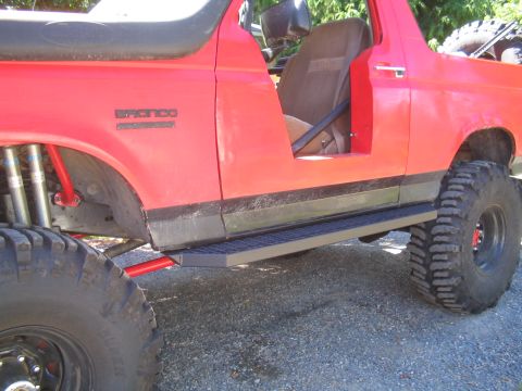 1989 Ford Bronco - A red 1989 Ford Bronco is shown from the side, equipped with large off-road tires and custom sliders.