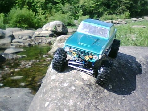 1996 Ford Bronco - A teal RC Bronco sits on a rock next to a shallow stream with trees in the background.