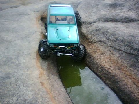 1996 Ford Bronco - This teal off-road RC car tackles a challenging rocky path with a water obstacle.