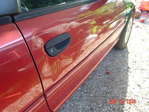 1997 Dodge Neon - A red car door shows significant damage, with a black door handle and a portion of the tire visible against a gravel background.