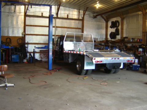 1992 Ford Bronco - Flatbed truck with dual rear wheels is parked inside a garage next to a car lift.