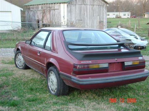 1984 Ford Mustang - Rear view of a maroon Ford Mustang with a spoiler and a "Mustang GT" badge, parked outdoors near buildings.