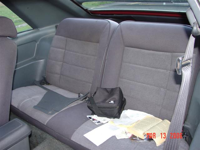 Back seat of a car with gray fabric seats, a black bag, and an open book on the seat. A watermark reads "MAR 13 2006 SUPERMOTORS.NET"