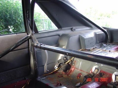 1984 Ford Mustang - The interior of a vehicle is shown, a prominent roll cage and a racing seatbelt, with visible rust on the floor pan.