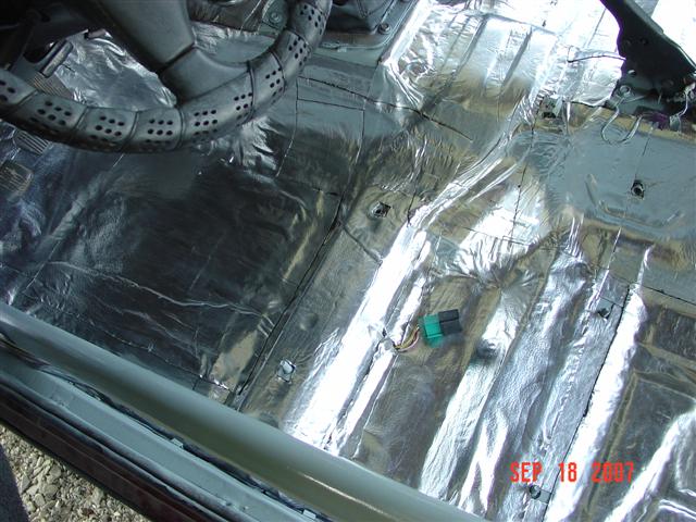 Car interior floor covered in silver sound deadening material, with part of the steering wheel visible
