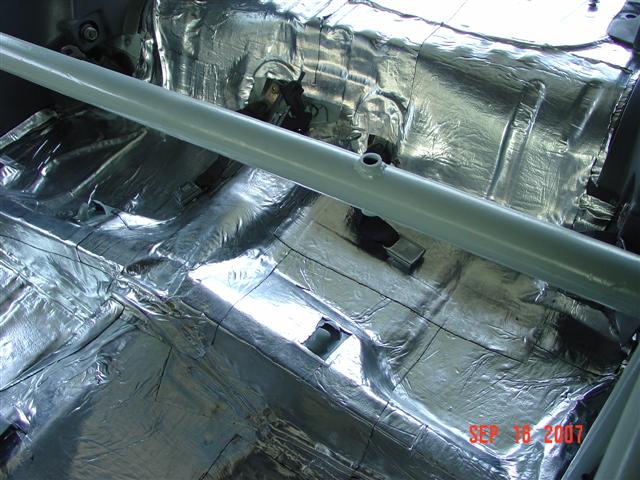 car interior floor covered in silver sound deadening material with a metal bar across the middle