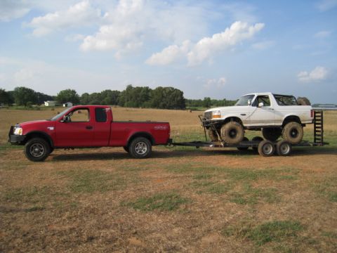 1996 Ford Bronco - Red Ford F-150 tows a modified white 1996 Ford Bronco on a trailer across a grassy field.