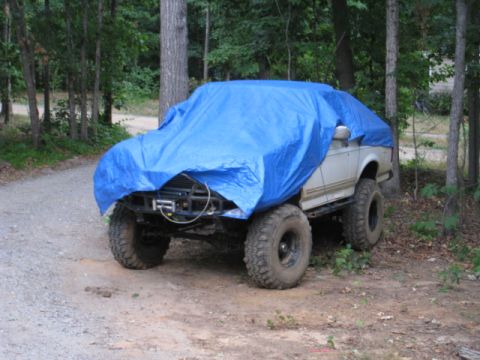 1996 Ford Bronco - A white SUV, partially covered by a blue tarp, sits on a dirt road surrounded by trees.