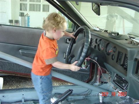 1984 Ford Mustang - A young child grips the steering wheel inside a vehicle's interior, with the dashboard and door panel in view.