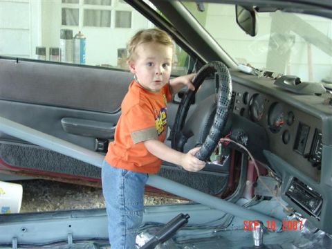 1984 Ford Mustang - Child sits in the driver's seat of a 1984 Ford Mustang, hands on the steering wheel.