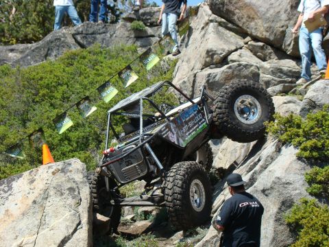 1989 Ford Bronco - A custom-built off-road buggy with large tires navigates a steep, rocky incline. Several small banners with logos hang on a line above the…