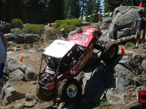 1989 Ford Bronco - A custom red off-road buggy with a white roof is navigating a rocky incline. The vehicle has large, knobby tires and a roll cage, with seve…