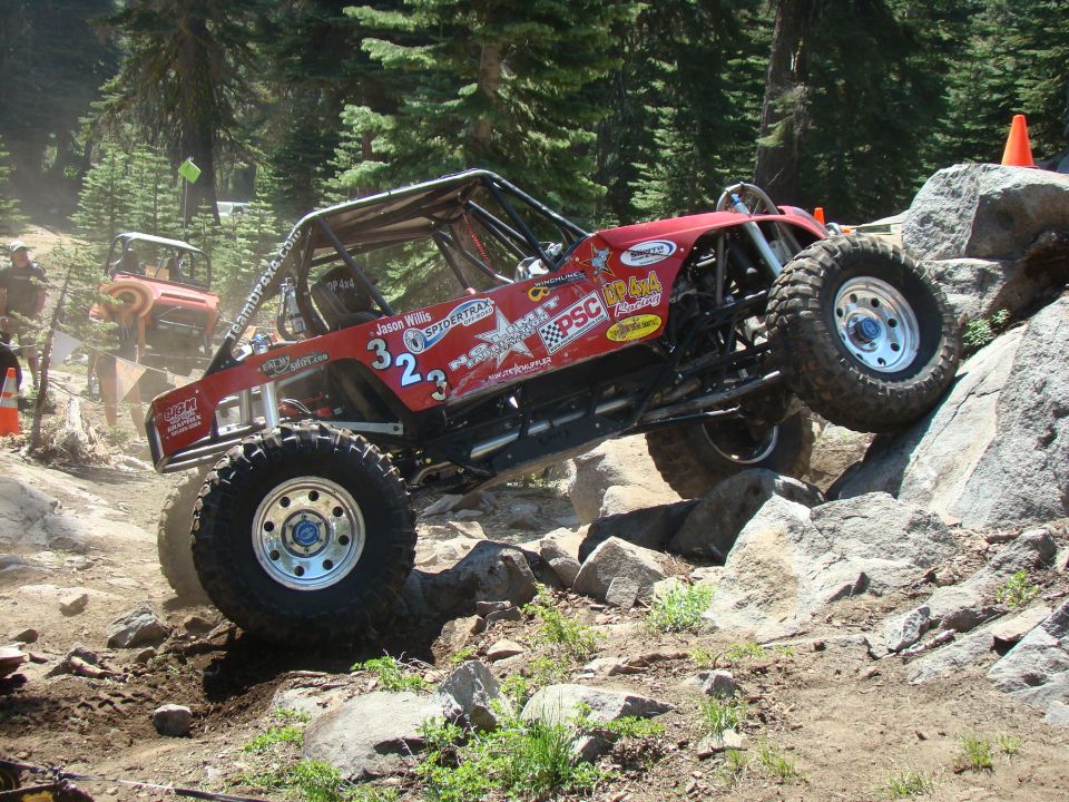 Red off-road buggy with large tires navigating a rocky incline. Another vehicle is visible in the background