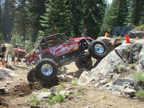 1989 Ford Bronco - A red off-road buggy with large tires navigates a rocky, wooded trail. The vehicle is tilted on its side as it climbs over large boulders.